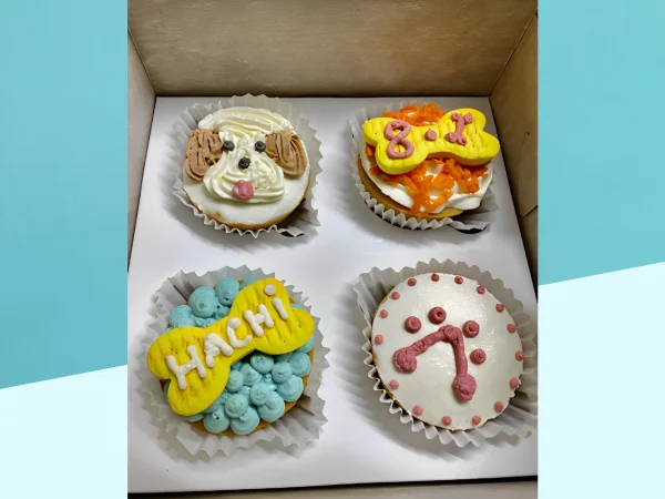 A set of four dog-themed cupcakes in a box, each decorated uniquely with colorful frosting and fondant elements.